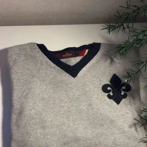 Grå v-ringad tröja från Morris - Snygg grå v-ringad tröja från Morris med mörkblå ribbad krage och två mörkblå ränder på ena ärmen. Stor dekorativ mörkblå lilja på bröstet. Tröjan är i mjuk bomull och har en klassisk, normal passform med långa ärmar.