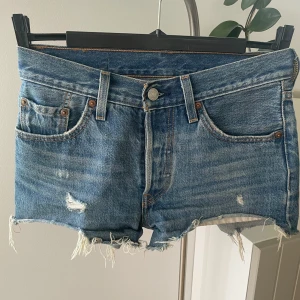 Levi's 501 jeansshorts  - Klassiska blå jeansshorts från Levi's 501 med råa, fransiga benslut och slitna detaljer. Femficksmodell med knappgylf och ikonisk läderpatch bak. Perfekta för en avslappnad och cool look under varma dagar.