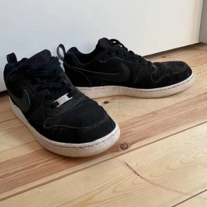 Svarta Nike Air Force 1 sneakers - Svarta Nike Air Force 1 sneakers med klassisk vit sula och svart swoosh på sidorna. Skorna har snörning och är tillverkade i mocka och syntet. Perfekta för dig som gillar en tidlös streetstil och vill ha ett par ikoniska sneakers i garderoben.