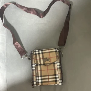 Rutig axelväska med brunt band från burberry - Snygg axelväska i klassiskt beige, svart och rött rutigt mönster. Väskan har ett brunt justerbart axelband, flera fack med dragkedja och en liten ficka framtill med metallspänne. Perfekt storlek för mobil, plånbok och småprylar.