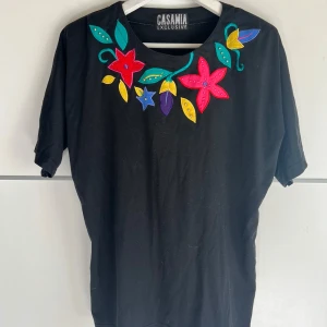 Svart blommig t-shirt från Casamia - Svart t-shirt från Casamia Exclusive med färgglada blommor och blad broderade vid halsringningen. T-shirten har korta ärmar och en klassisk passform. Perfekt för dig som vill sticka ut med en unik och färgglad detalj.