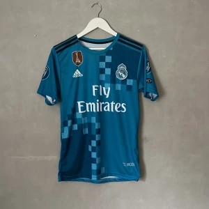 Real madrid 2017/2018 - Säljer en officiell Real Madrid Champions League-tröja från Adidas med Ronaldo 7 på ryggen. Tröjan är blå med pixelmönster och har tryckta märken som Fly Emirates och CL 2017 på ärmen. Kortärmad och tillverkad i lätt, ventilerande material.