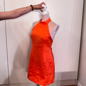 Orange halterneck kortklänning - Snygg orange kortklänning med halterneck och öppen rygg. Klänningen har en figurnära passform och är tillverkad i ett glansigt material som ger en cool look. Perfekt för dig som vill sticka ut med färg och stil.