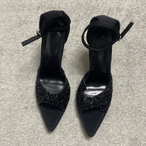 Svarta glittriga pumps med klack - Snygga svarta pumps med spetsig tå och öppen tå. Skorna har glittriga detaljer framtill och en smal klack. Rem runt ankeln med spänne för extra support. Perfekta för dig som vill sticka ut lite extra.