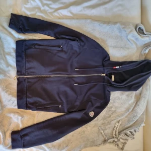Mörkblå Moncler hoodie med dragkedja - Snygg mörkblå hoodie från Moncler med hel dragkedja, huva med snörning och två fickor med dragkedja. Klassisk Moncler-logga på ärmen. Tillverkad i mjuk bomull, perfekt för chill dagar eller när du vill ha en clean look. Haft koftan en längre tid men säljer nu då den blivit för liten, den är i mycket bra skick. 