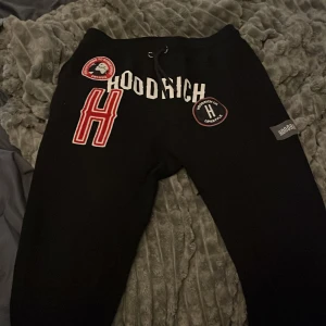 Svarta mjukisbyxor från Hoodrich S - Svarta mjukisbyxor från Hoodrich med coola patches och broderade detaljer framtill. Byxorna har snörning i midjan och flera tryck, bland annat ett stort rött H och Hoodrich-logga. Perfekta för en avslappnad streetwear-look.