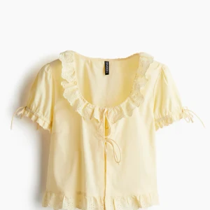 Gul blus med volang och knyt från Divided - Supersöt ljusgul blus från Divided med volangkant runt halsen och nederdelen, samt knyt framtill. Blusen har korta puffärmar med knyt och broderade detaljer på volangerna. Perfekt för dig som gillar romantisk och feminin stil.