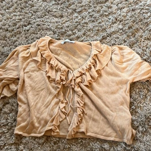 Aprikos stickad bolerotopp från Zara - Supersöt beige stickad kofta från Zara med volangkant längs ringningen och framtill. Toppen har korta puffärmar och knytning framtill. Man kan både ha med och utan linne under. Perfekt till allt.