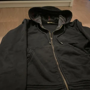 Svart hoodie från Burberry med dragkedja - Svart hoodie från Burberry med hel dragkedja och klassisk huva. Insidan av huvan har rutigt mönster i grått och rött. Tröjan är i mjuk bomull och har fickor framtill. Perfekt för dig som gillar stilrena och exklusiva plagg.