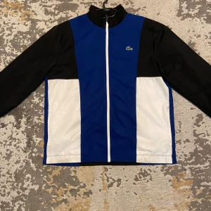 Lacoste Tracksuit Storlek L - Otroligt snygg och bekväm Lacoste Tracksuit. Stort krokodil märke och texten ”Lacoste” på baksidan med ett mindre märke på framsidan av koftan. Byxor medföljer! Svarta byxor med en vit linje längs byxornas sidor och ett Lacoste märke vid midjan. Köptes i Barkarby Outlet i Stockholm, Lacoste affär, säljes för 1000kr. Pris kan diskuteras! Passar storlek L. Det finns tecken på användning på tracksuiten. Den är alltså inte helt ny! 
