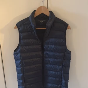 Mörkblå dunväst från Uniqlo S - Säljer en mörkblå, ultralätt dunväst från Uniqlo i storlek S. Västen har horisontella quiltade sömmar, hög krage och dragkedja framtill. Perfekt att slänga över en hoodie eller tröja när det är kyligt ute.