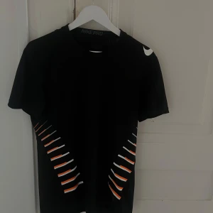 Svart Nike Pro t-shirt med tryck - Svart t-shirt från Nike Pro med coolt grafiskt tryck i vitt och orange längs sidorna. Klassisk rund hals och korta ärmar. Perfekt för dig som gillar sportig stil och vill sticka ut lite extra. Snygg Nike-logga på axeln.