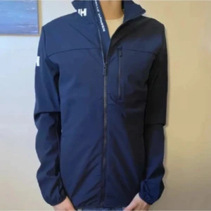 Blå vindjacka från Helly Hansen - Snygg marinblå vindjacka från Helly Hansen med dragkedja hela vägen, hög krage och logga på axeln. Jackan har en bröstficka med dragkedja och är perfekt för blåsiga dagar. Enkel och stilren design som funkar till det mesta.