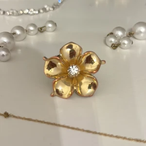 Guldig blomring Ioaqu - Ringen är i Ok skick har rostat på baksidan, syns inte när man har på sig den. Vänligen se bilder. Säljer en snygg ring i form av en blomma med fem blad i guldton och en glittrande kristall i mitten. Ringen har öppen baksida så storleken kan justeras. Perfekt för dig som gillar unika och blommiga detaljer.