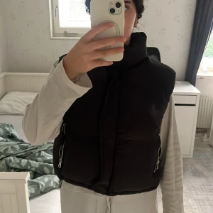 Svart dunväst från FB Sister - Svart, quiltad dunväst från FB Sister med hög krage och två fickor med dragkedja. Västen har en clean look och är perfekt att slänga över en hoodie eller långärmad tröja. Croppad modell som ger en trendig siluett. 