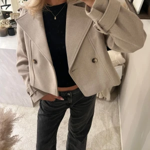 Beige cropped trenchcoat kappa från Asos Design - Snygg croppad trenchcoat kappa i beige med breda slag, dubbelknäppning och knappar vid ärmslut. Jackan har axelklaffar och är tillverkad i ett ull-liknande material som ger en exklusiv känsla. Perfekt för dig som vill ha en stilren och trendig look. Nypris 799kr och aldrig använd!!🤎🤎