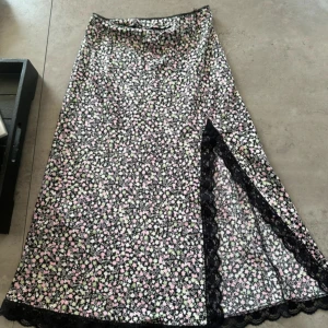 Blommig långkjol med spets från H&M Divided - Supersöt långkjol från H&M Divided med svart botten och små blommor i rosa, vitt och grönt. Kjolen har en slits med svart spetskant som ger en extra detalj. Tillverkad i mjuk polyester med lite stretch för skön passform.