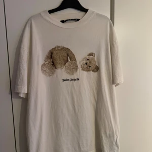 Palm Angels vit t-shirt med björntryck - Palm Angels t-shirt i vitt med stort tryck av en sönderdelad nallebjörn på framsidan och Palm Angels-logga i svart på både fram- och baksidan. T-shirten har en loose passform, rund hals och är tillverkad i mjuk bomull. Perfekt statement-plagg för dig som gillar streetwear.