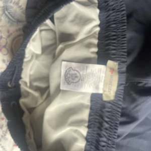 Mörkblåa badshorts från Moncler - Snygga svarta badshorts från Moncler med elastisk midja och snörning. Shortsen har en bakficka med knapp och Moncler-logga framtill. Perfekta för stranden eller poolen, klassisk design och riktigt sköna att bära. Storlek M passar S. Original moncler lappar finns med.
