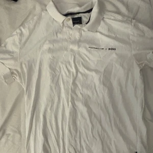 Vit Porsche BOSS pikétröja - Vit pikétröja från Porsche x BOSS med klassisk krage och knappslå. Diskret logga broderad på bröstet. Perfekt för dig som gillar stilren design och vill ha en t-shirt med lite extra attityd. Skön bomullskvalitet och korta ärmar.