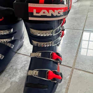 Mörkblå Lange RSJ 60 pjäxor - Säljer ett par mörkblå Lange RSJ 60 pjäxor med röda detaljer och fyra metallspännen för perfekt passform. Snygg och sportig design med tydlig logga och robust yttersula. Perfekta för dig som vill ha stilrena och funktionella pjäxor i backen. storlek 249 mm