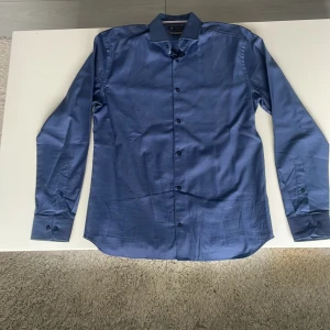 Blå slim fit skjorta från Riley - Snygg blå skjorta från Riley i slim fit-modell. Skjortan har klassisk krage, långa ärmar med manschett och knappar framtill. Perfekt för dig som gillar en stilren look med modern passform. Perket skick