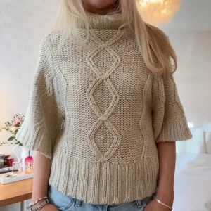 Beige stickad polotröja med kort ärm - Säljer en chunky beige stickad polotröja med korta ärmar och snyggt kabelstickat mönster framtill. Tröjan har bred ribbad mudd i nederkant och vid ärmslut, perfekt för lager-på-lager och chill höststil.