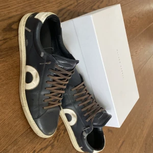 Oliver Cabell Low 1 Oxford sneakers - Snygga Oliver Cabell Low 1 sneakers i mörkblått skinn med vita detaljer och bruna snören. Klassisk låg modell med rund tå och platt sula. Stora vita cirklar på sidorna ger en unik look. Perfekta för dig som gillar stilrena men ändå unika sneakers. Priset inte hugget i sten vill helst bli av med dem. 