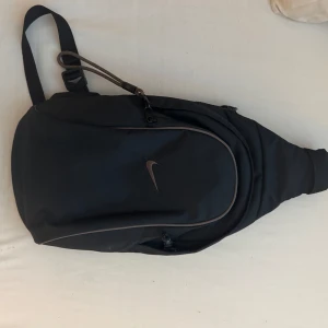  Nike - Snygg svart ryggsäck från Nike med minimalistisk design och diskret logga på framsidan. Väskan har dragkedja, justerbar rem och är tillverkad i slitstarkt syntetmaterial. Perfekt för att bära dina grejer på ett stilrent sätt.  Sling bag!   Axelväska!     