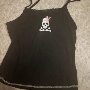 Svart linnetopp med döskalle från Romwe - Svart linnetopp från Romwe med tunna axelband och spetskant upptill. På bröstet finns ett broderat döskallemotiv med en rosa rosett. Perfekt för dig som gillar edgy detaljer och vill sticka ut lite extra.