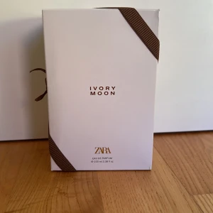Zara Ivory Moon Eau de Parfum 100ml - Snygg parfym från Zara, Ivory Moon, i en stilren vit kartong med bruna banddetaljer. Flaskan rymmer 100 ml Eau de Parfum och har en modern, minimalistisk design som känns lyxig och trendig. Perfekt för dig som gillar eleganta och fräscha dofter.
