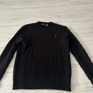 Svart kabelstickad tröja Polo Ralph Lauren - Kabelstickad svart tröja från Polo Ralph Lauren med klassisk rund hals och röd broderad logga på bröstet. Tröjan har lång ärm och ribbade muddar. Perfekt för dig som gillar stilren och tidlös design.