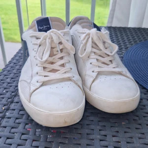 Vita sneakers från Pepe Jeans  - Snygga vita sneakers från Pepe Jeans med marinblå och beige detaljer. Klassisk modell med snörning, rund tå och platt sula. Skorna har logga i guld på sidan och '1973' präglat bak på sulan. Ovandel i skinn och textil för en fräsch look.