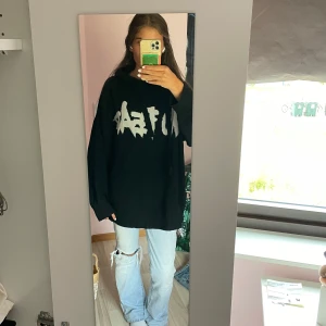 Svart hoodie med vit text - Säljer en oversized svart stickad hoodie med vit grafisk text framtill. Tröjan har luva och långa ärmar med breda muddar. Materialet är stickat och känns riktigt mjukt och skönt. Perfekt för dig som gillar streetstyle och vill ha en chill look.