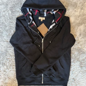 Burberry Zip Up Hoodie  - Lyxig hoodie från Burberry i svart med den klassiska rutiga huvan som detalj. Ikonisk och tidlös design med högsta kvalitet – perfekt för både vardag och mer dressade tillfällen.  ♦️ Helt ny – med originaltag kvar ☑️ ⚜️ ☑️ ♦️ Exklusiv Burberry-detalj i huvan ☑️  📦 Snabb leverans & trygg affär 
