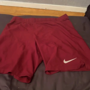 Vinröda Nike shorts med logga - Snygga vinröda shorts från Nike med klassisk vit logga på benet. De har en loose passform och är tillverkade i ett lätt syntetmaterial som passar perfekt för träning eller sport. Elastisk midja för skön komfort och rörelsefrihet.