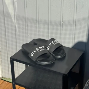 Svarta sandaler från Givenchy - Svarta slides från Givenchy med vit logotyp på bred rem över foten. Klassisk och stilren design, perfekt för sommardagar eller chill hemma. Tillverkade i syntetmaterial med platt sula och öppen tå.