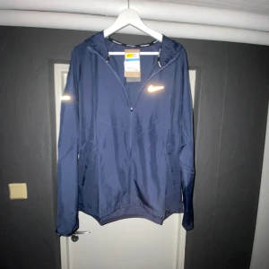 Blå vindjacka från Nike med huva - Snygg marinblå vindjacka från Nike med reflekterande logga på bröstet och detaljer på ärmarna. Jackan har huva, dragkedja framtill och två fickor. Perfekt för dig som gillar sportig stil och vill ha något lätt och smidigt till vardagen.