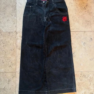 Svart lång jeanskjol från JNCO Jeans - Grym svart lång jeanskjol från JNCO Jeans med kontrastsömmar och stora fickor. Snyggt broderi i rött och färgglatt motiv på bakfickan ger kjolen en unik streetkänsla. Klassisk femficksmodell med dragkedja och bälteshällor.