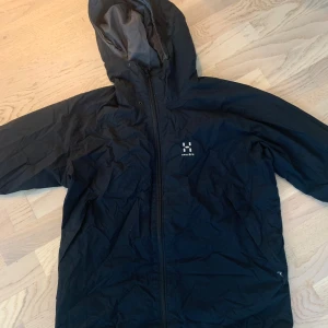 Svart vindjacka från Haglöfs - Svart vindjacka från Haglöfs med huva och hel dragkedja framtill. Jackan har en stilren design med diskret logga på bröstet och meshfoder på insidan för extra ventilation. Perfekt för blåsiga dagar och outdoor-äventyr. —— moncler väst 2 för 1 