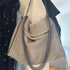 Grå axelväska med kedjedetaljer - Snygg grå axelväska i mocka-look med kraftiga silverfärgade kedjor som axelrem och runt kanterna. Väskan har en mjuk och slouchy form, perfekt för dig som vill ha en cool och trendig accessoar till din outfit.