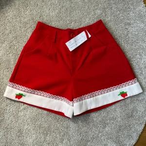 Röda shorts med spets och jordgubbar - Superfina röda shorts med vit kant nertill, dekorerad med spets och broderade jordgubbar på båda benen. Modellen har hög midja, fickor och en röd knapp framtill. Perfekt för dig som vill sticka ut med en söt och färgglad look. Kawaii och köpt från Japan!