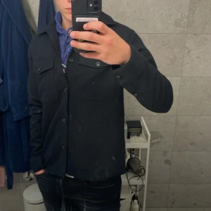 Svart overshirt från J.Lindeberg - Svart overshirt från J.Lindeberg i storlek M. Overshirten har klassisk krage, två bröstfickor med knappar och långa ärmar med manschettknappar. Snygg och stilren design som funkar till många olika looks. Nästan inte använd! 
