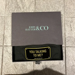 Grön mössa John Hatter & Co - Svart mössa från John Hatter & Co med uppvikt kant och en metallskylt framtill där det står 'YOU TALKING TO ME?'. Skylten har guldfärgade detaljer som ger en cool och edgy vibe. Perfekt för dig som vill sticka ut med din stil.