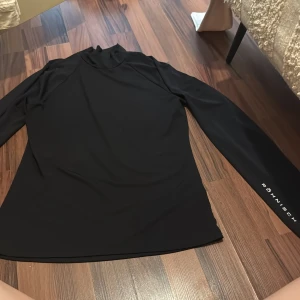 Svart långärmad träningströja Röhnisch - Svart långärmad träningströja från Röhnisch med diskret logga på ärmen. Tröjan har en hög krage och är gjord i mjukt, stretchigt syntetmaterial som andas. Perfekt för gymmet eller löprundan när du vill ha en clean och sportig look.