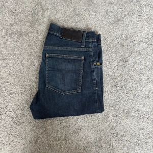 Mörkblå jeans Iggy 32/34 - Säljer ett par mörkblå jeans från tiger of Sweden i modell iggy Klassisk femficksdesign, raka ben och snygg tvätt. Jeansen är tillverkade i bomull och har en stilren look som passar till det mesta.