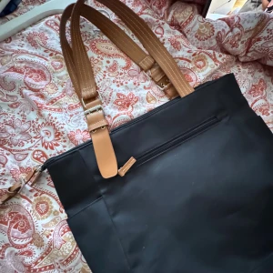 Svart axelväska med bruna detaljer! - Stilren svart axelväska med rejäla bruna axelband i skinnimitation. Väskan har dragkedja upptill, ytterfack med dragkedja och rymligt huvudfack. Perfekt för dig som vill ha plats för allt du behöver under dagen.