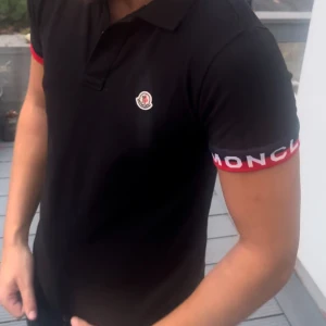 Svart Moncler pike med röd detalj - Svart t-shirt från Moncler med korta ärmar och röd kant med logotyp på ärmsluten. Klassisk krage och liten Moncler-logga på bröstet. Perfekt för dig som gillar stilrena märkesplagg med snygga detaljer.