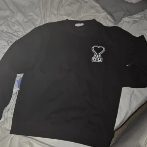 Svart sweatshirt från Ami paris S - Svart sweatshirt från Ami i bomull med ikonisk vit broderad hjärtlogo på bröstet. Klassisk rund halsringning och långa ärmar. Perfekt för en clean och stilren look, passar till jeans eller sweatpants. Sitter som s