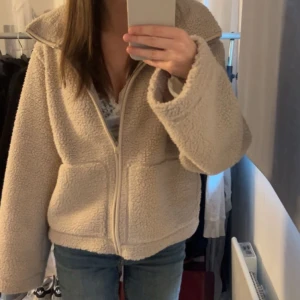 Beige teddyjacka med dragkedja - Mysig beige teddyjacka med dragkedja framtill och stora fickor. Jackan har en hög krage och är perfekt för kyliga dagar. Den har en loose passform och är tillverkad i ett fluffigt teddy-material som känns supermjukt.
